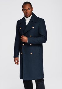 Marineblaue Wolle Doppelreiher mit großen goldenen Knöpfen, breitem Kragen und Seitentaschen. Getragen über einem weißen Rollkragenpullover.
