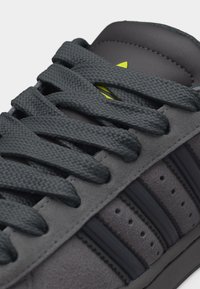 Εγγύς πλάνο γκρι αθλητικού παπουτσιού με υφασμάτινα σκούρα γκρι κορδόνια, μαύρες ρίγες στις πλευρές σουέντ, και ένα μικρό εμφανές κίτρινο λογότυπο κάτω από τα κορδόνια.