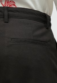 Sötétszürke nadrág sima felülettel, övhurkokkal, lapos elejével és egyetlen hátsó zsebével, minimális varrási részletekkel.