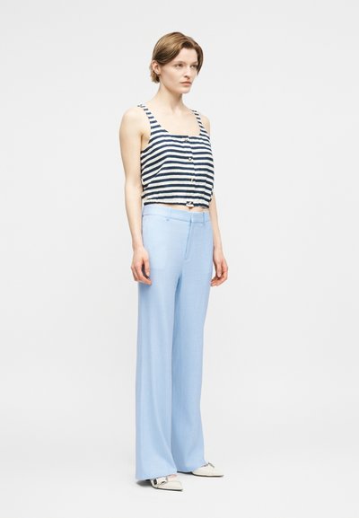 Γυναίκα που στέκεται φορώντας μπλε σκούρο και λευκό ριγέ crop top χωρίς μανίκια με κουμπιά, ανοιχτό μπλε παντελόνι με ευρύ πόδι και λευκά μυτερά παπούτσια.