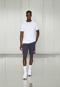 Bela majica z okroglim izrezom, mornarsko modre kratke hlače z logotipom, bele nogavice in športni čevlji. Model stoji na odsevnem tleh pred kovinskim ozadjem.