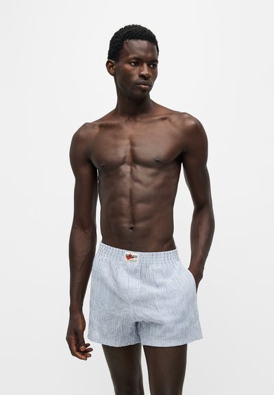 Homme musclé à la peau foncée portant un short rayé bleu clair et blanc, debout devant un fond clair uni.