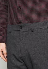 Camicia a quadri con bottoni in bordeaux e nero, abbinata a pantaloni grigi su misura. I pantaloni presentano una chiusura con un solo bottone e un design a fronte piatta.