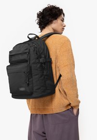 Black backpack with multiple zippered pockets, textured fabric, and adjustable straps, worn over a cozy orange sweater.