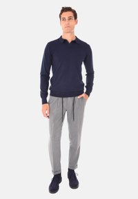 Maglione blu navy con colletto standard, abbinato a pantaloni a pinces grigi chiari con vita regolabile e scarpe scure.