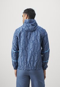 Giacca a vento blu con cappuccio, caratterizzata da un motivo numerico testurizzato, orlo elastico e strisce blu lungo le maniche. Materiale leggero.