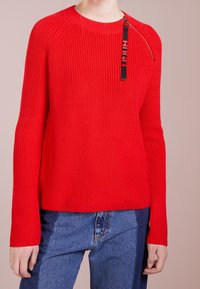 Punainen neulottu neulepaita, jossa on resorikuvio, pitkät hihat ja kauluksessa vetoketju, jossa on merkitty vetotappi. Yhdistetty sinisiin farkkuihin.