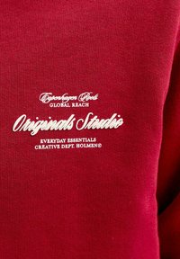 Tissu rouge avec texte imprimé en blanc mentionnant "Originals Studio" et des phrases plus petites incluant "Copenhagen Root", "Global Reach" et "Everyday Essentials Creative Dept. Holmen".