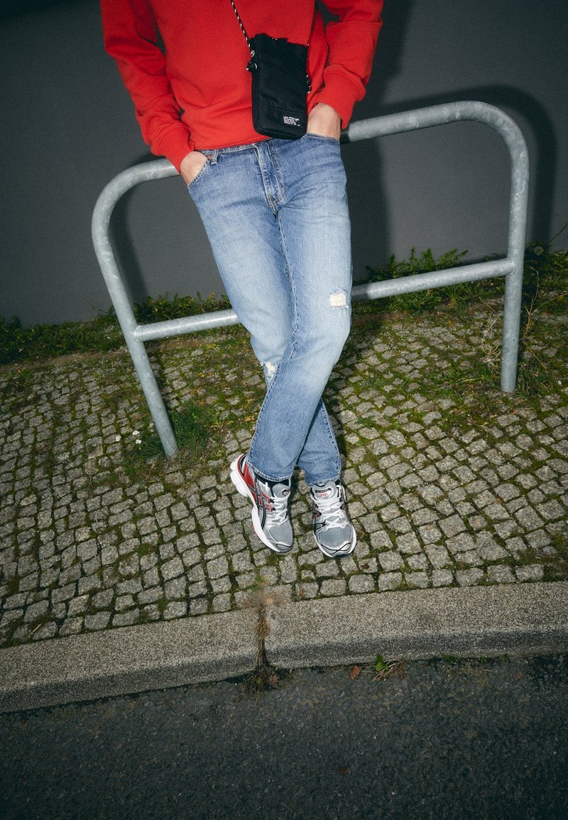 Un jean en denim bleu avec des détails déchirés, associé à des baskets de sport grises et rouges, et un sac bandoulière noir sur un sweat-shirt rouge.