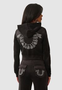 Hoodie en suédine noire avec capuche, présentant des embellissements en strass en motif courbé à l'arrière. Pantalons taille haute assortis avec poches.