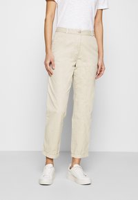 Chino beige con taglio dritto, leggermente accorciati alle caviglie, dotati di chiusura con bottone, tasche laterali e orli risvoltati. Indossati con sneakers bianche.
