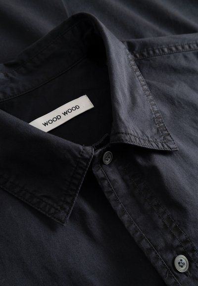 Tumesinise nuppudega särgi krae lähivõte, mille sees on valge silt tekstiga "WOOD WOOD" õmmeldud kaeluse siseküljele.