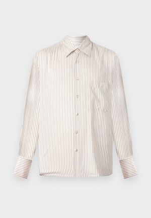 Νέος άντρας όρθιος με τα χέρια στις τσέπες, φοράει μπεζ ριγέ μακρυμάνικο πουκάμισο, μαύρο παντελόνι με φαρδιά πόδια και μαύρα παπούτσια σε λευκό φόντο.