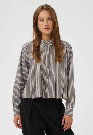 Jeune femme aux longs cheveux bruns portant une chemise à rayures verticales grises et blanches et un pantalon noir, debout devant un fond uni.