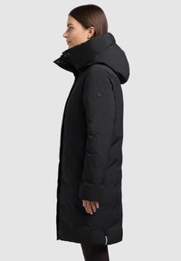 Melna polsterēta jaka ar augstu apkakli un kapuci, sānu kabatām un plānu siluetu. Gluda tekstūra ar minimālām detaļām.