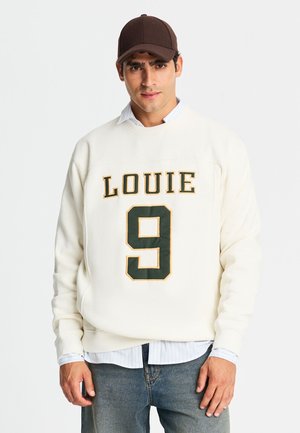Uomo che indossa una felpa bianca con "LOUIE 9" in verde e giallo, un berretto marrone, una camicia azzurra e jeans blu, in piedi davanti a uno sfondo semplice.
