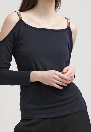 Femme portant un top noir épaules dénudées avec des détails en lanières de cuir, debout les mains jointes devant elle.