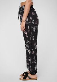 Femme debout de côté portant un pantalon noir à imprimé floral avec des poignets élastiques, un crop top blanc, des sandales noires et un bracelet �à charms.