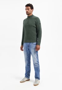 Groene geribbelde trui met een hoge kraag, gecombineerd met lichtblauwe denimjeans en grijze sneakers met witte strepen.