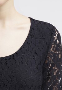 Schwarzes Spitzentop mit floralem Muster, featuring einem schmalen Rundhalsausschnitt und halbtransparenten langen Ärmeln. Glatte Stoffstruktur, figurbetontes Design.
