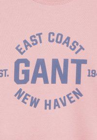 Jasnoróżowa bluza z okrągłym dekoltem z ciemnoniebieskim nadrukiem tekstowym: "EAST COAST GANT EST. 1949 NEW HAVEN." Gładka bawełniana tekstura.