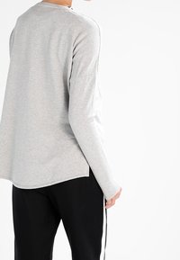 Persoon die een lichtgrijs shirt met lange mouwen en een zwarte broek met witte zijstrepen draagt, gezien van achteren tegen een witte achtergrond.