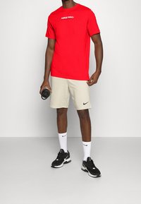 Punainen Nike Pro -t-paita, beiget shortsit, mustat Nike-kengät, valkoiset sukat logolla. Pitää mustaa vesipulloa, seisoo neutraalilla taustalla.