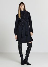 Trench coat in misto lana nera con bottoni doppi, vita cinturata e tasche a patta. Il modello indossa stivali di pelle nera al ginocchio.