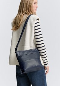 Borsa a tracolla in pelle blu navy con superficie testurizzata, tracolla regolabile e dettaglio con tasca con zip. Indossata con un gilet bianco e una maglietta a righe.