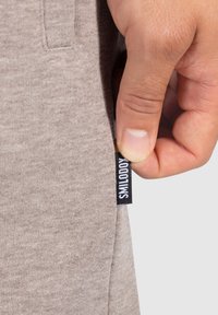 Tejido textil beige siendo sostenido, con una etiqueta que dice "SMILODON" en blanco sobre un fondo negro, y sutiles detalles de textura visibles.