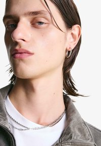 Primo piano di una persona con capelli castani bagnati che indossa un orecchino a cerchio argentato, collane di argento sovrapposte, una camicia bianca e una giacca grigia su sfondo bianco.