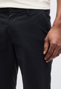 Gros plan d'une personne portant un pantalon noir et une chemise blanche, la main droite reposant le long du corps.
