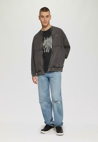 Grijze zip-up jas met ribgebreide manchetten over een zwart graphic T-shirt, gecombineerd met lichtblauwe versleten jeans en zwarte sneakers.