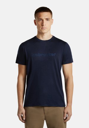 Männliches Model trägt ein marineblaues T-Shirt mit Rundhalsausschnitt und dem Text "icebreaker" sowie braune Hosen, steht vor einem einfarbigen hellen Hintergrund.