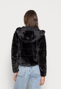 Giacca corta in pelliccia sintetica nera con cappuccio, caratterizzata da una superficie testurizzata e dettagli di cuciture orizzontali, abbinata a jeans azzurri.