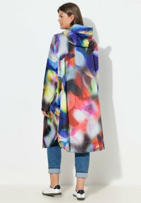 Impermeable colorido y ligero con capucha y un patrón floral abstracto. Cuenta con mangas largas y cierres de botones. Se lleva sobre jeans cortos y zapatillas.