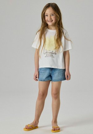 Ung pige smiler, iført hvid t-shirt med grafik, blå denimshorts og gule klipklappere, stående mod en ensartet lys baggrund.