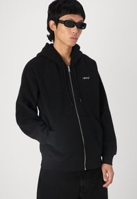 Hanorac negru din fleece, cu fermoar, dotat cu un buzunar frontal, glugă cu șnur și un logo branduit pe piept. Mânecile cu tiv din tricot completează designul.