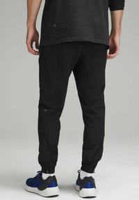 Schwarze Jogginghosen verfügen über ein schmal zulaufendes Design mit elastischen Bündchen, einer glatten Textur und seitlichen Reißverschlusstaschen. Kombiniert mit blauen Sportschuhen.