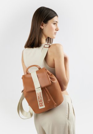 Giovane donna con capelli castani lisci che indossa un top beige senza maniche e pantaloni, con uno zaino in pelle marrone con tracolla bianca e dettaglio testo.