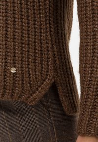 Pull en tricot marron avec une texture côtelée, doté d'un d�étail de bouton en argent et d'un ourlet fendu. Porté par-dessus un tissu à carreaux.