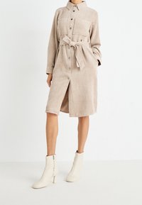 Vestito camicia in corduroy beige con bottoni frontali, due tasche sul petto, cintura annodata in vita e lunghezza fino al polpaccio. Abbinato a stivaletti bianchi alla caviglia.