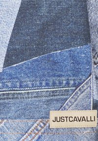 Denimtyg med blå nyanser i olika toner, med en sydd etikett som står "JUSTCAVALLI." Texturerad med distinkta sömmar och mönster.