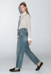 Leicht gestreiftes Button-up-Hemd mit weißem Kragen, kombiniert mit hellen, straight-leg Jeans und schwarzen Slippers. Glatte Textur und lässige Passform.