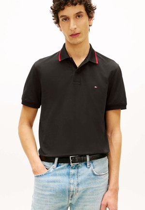 Jeune homme portant un polo noir avec une bordure rouge et un jean bleu clair, debout avec une main dans la poche sur fond blanc.