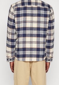 Camicia in flanella a quadri con colori blu, crema e beige, maniche con doppio bottone e design leggermente accorciato. Tessuto morbido e texturizzato.