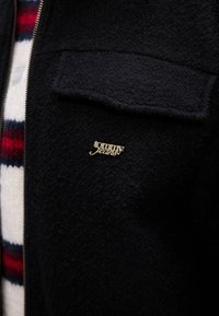 Sort fleecejakke med lynlås, der har en lappelomme og et guld "tommy jeans" logo på brystet. Materialet har en tekstureret overflade.