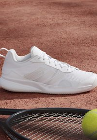 Scarpa sportiva bianca su un campo da tennis in terra battuta accanto a una racchetta da tennis nera e una palla da tennis gialla.