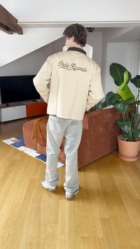 Beige Jacke mit braunem Kragen, "Indie Records" auf dem Rücken gestickt, kombiniert mit hellblauen Jeans und silbernen Sneakers in einem Wohnzimmer.