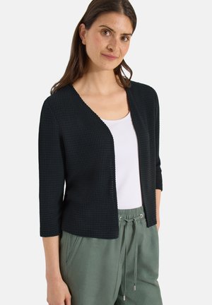 Kobieta w czarnym teksturowanym kardiganie z otwartym przodem, na białym topie i zielonych spodniach ze sznurkiem, stojąca na jednolitym tle.